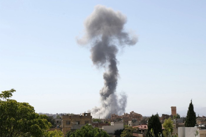 A plume of smoke rises high into the air from a town where there are red roofed houses and a belltower