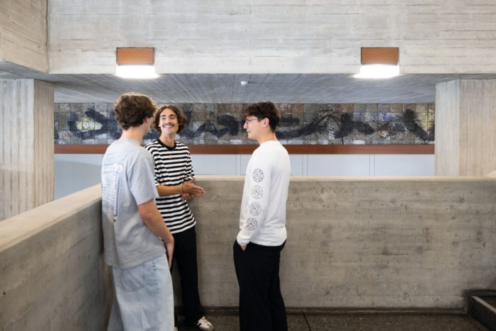 Three casually dressed young men engage in conversation in a modern building with exposed concrete walls and an abstract mural in the background