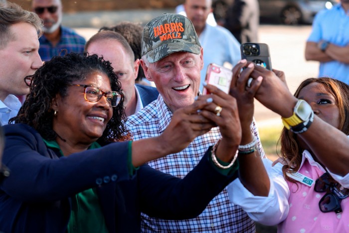 Bill Clinton, wearing a cap with ‘Harris Walz’, takes selfies with enthusiastic supporters holding up phones.