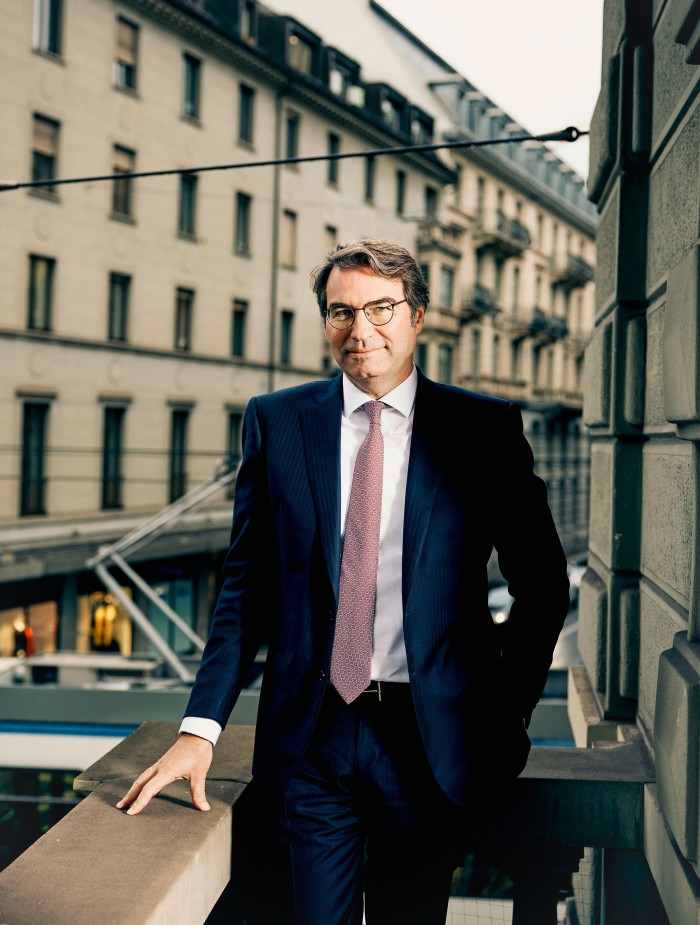 A man stands on a balcony or terrace, dressed in a dark, tailored suit with one hand resting casually on the railing