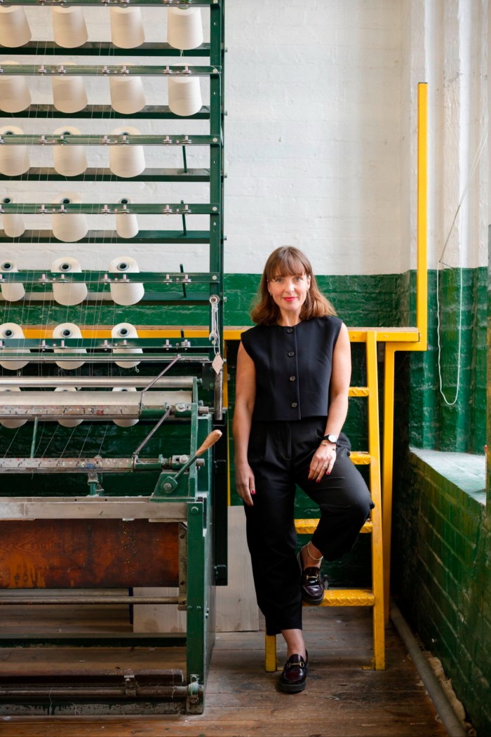 A woman dressed in black leans against a yellow metal ladder beside a lace loom