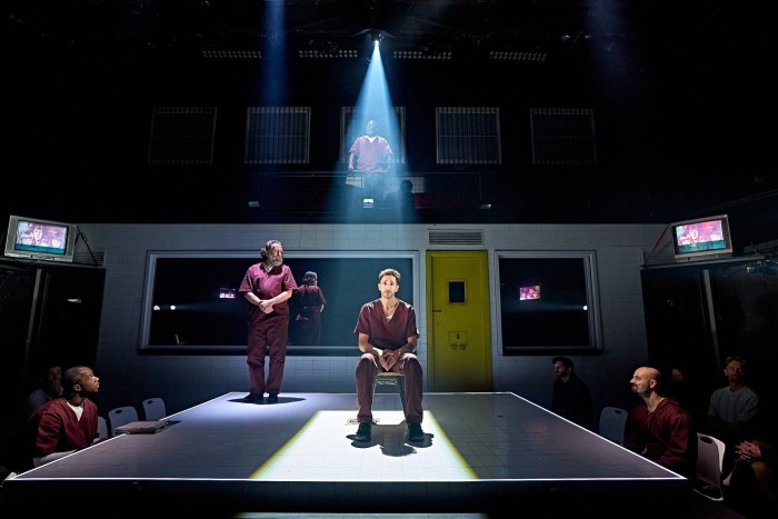 A man in a purple prison uniform is seated under a spotlight while other inmates look on