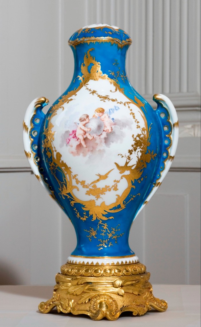 Porcelain vase with a gilded base, coloured royal blue with a white diamond in the middle which has gold-leaf frame and features a painting of two cherubs in the centre