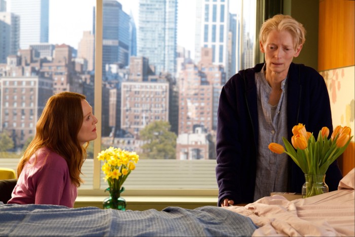 In a room with a large window looking out on to New York skyscrapers, a woman sits next to a bed looking up at another woman wearing a dressing gown; she stands, looking ill and anxious. There are two vases of flowers