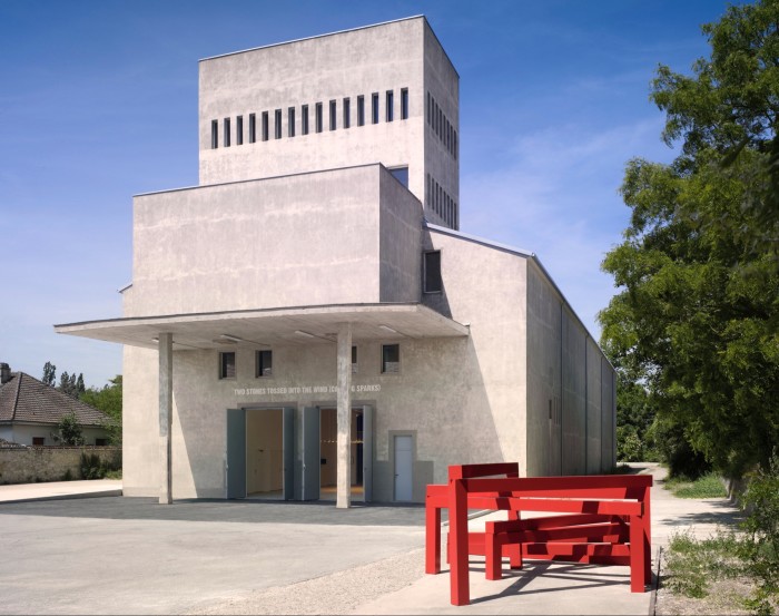 Photograph of an unusually shaped, white-walled modernist building, with two sets of doors open at the front, under a flat shelter, and a quote above the doors reading “Two stones tossed into the wind (causing sparks)”