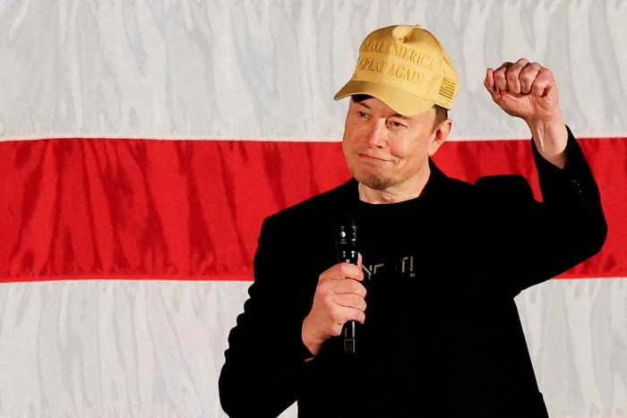 Elon Musk, wearing a &quot;Make America Great Again&quot; hat, gestures and holds a microphone while speaking in front of a backdrop with red and white stripes.