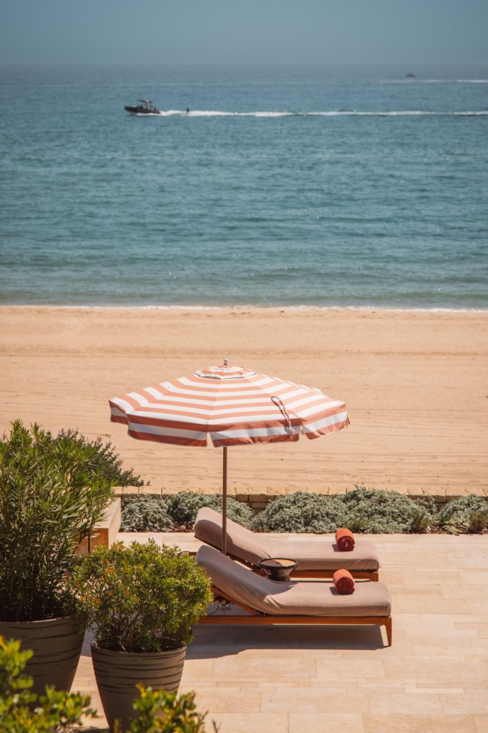 Two sun loungers, partially shaded by a striped parasol above them, next to a sandy beach, with a speedboat visible in the background