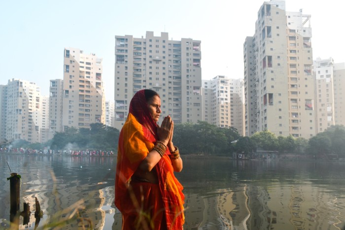 A woman dressed in a traditional red and orange sari stands with her hands in a prayer position by a reflective body of water, with high-rise buildings and greenery in the background