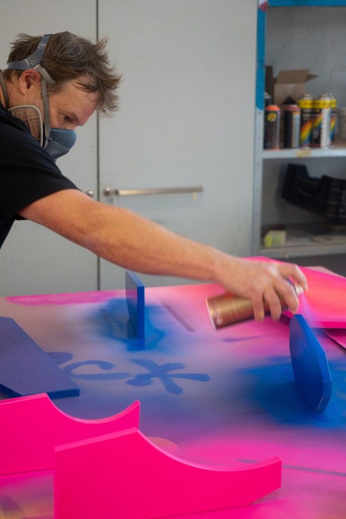 A man with a respirator mask is holding a spray paint can in his right hand, applying a layer of paint onto various cut-out shapes on a work surface