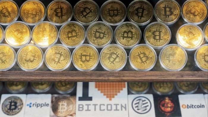 Rows of gold-coloured bitcoin tokens displayed in a window, with signage below showcasing various cryptocurrency logos