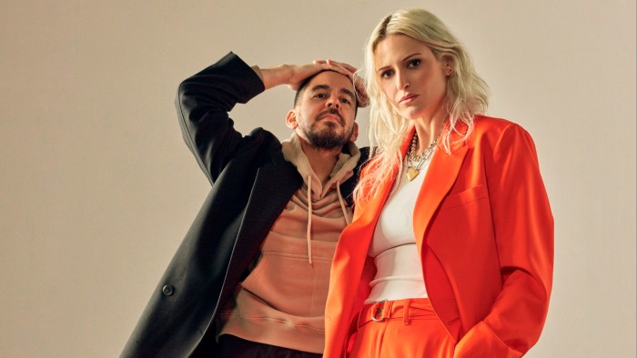 A man in a beige hoodie and black jacket poses with his hands on his forehead next to a blond woman in a bright orange suit