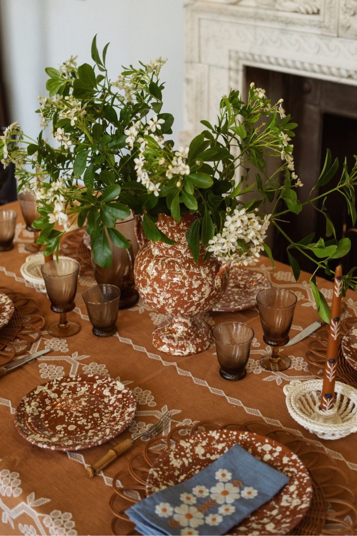 A table set with brown glasses and ceramic plates, which are brown with an off-white splatter pattern. A jug in the same design is filled with white hydrangeas. There is a pale brown embroidered tablecloth, and a blue embroidered napkin sits folded on one of the plates.