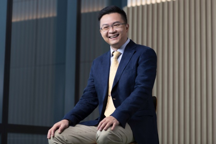 A man in formal attire, including a yellow tie and navy jacket, sitting and smiling warmly