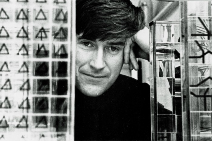 A black and white, head and shoulder shot of a man with dark hair. which light boxes on either side of him. He is smiling, and leaning his head in his hands