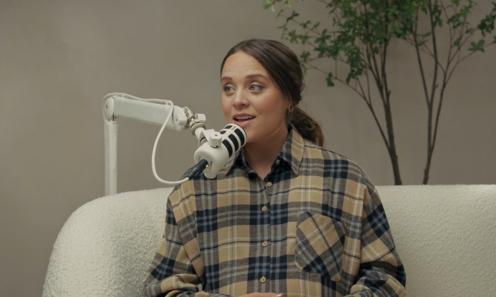 Jinger Duggar speaks into a mic while wearing a beige plaid shirt.