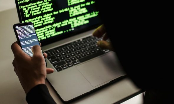 A hacker typing on a MacBook laptop with code on the screen.