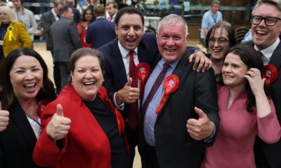 Davy Russell, Anas Sarwar, Jackie Baillie, and Monica Lennon celebrate together at South Lanarkshire Council Headquarters in Hamilton, Scotland. They are smiling and giving thumbs up, surrounded by supporters.