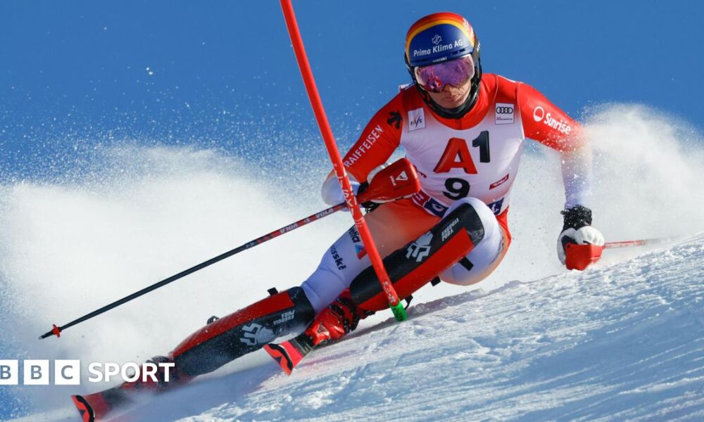 Alpine skier in red and white racing suit carving through snow on a steep slope, leaning sharply with poles extended, snow spraying behind against a clear blue sky.
