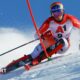 Alpine skier in red and white racing suit carving through snow on a steep slope, leaning sharply with poles extended, snow spraying behind against a clear blue sky.
