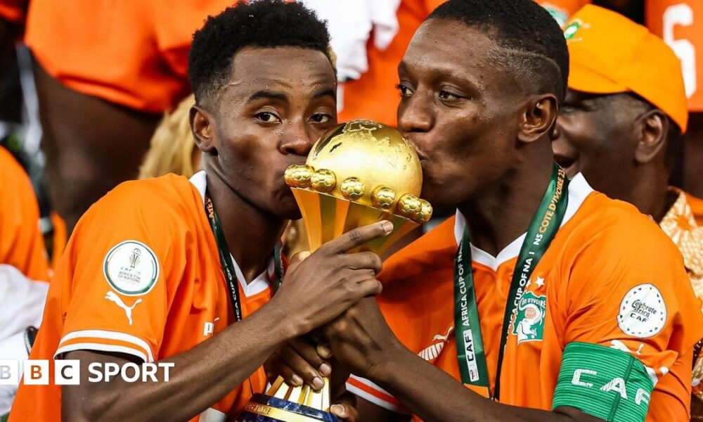 Simon Adingra and Max-Alain Gradel seen from chest up, both wearing bright orange Ivory Coast football shirts with medals around their necks, hold the golden Africa Cup of Nations trophy between them and kiss the top of it. Adingra is looking directly into the camera while Gradel casts his eyes to the left of the screen