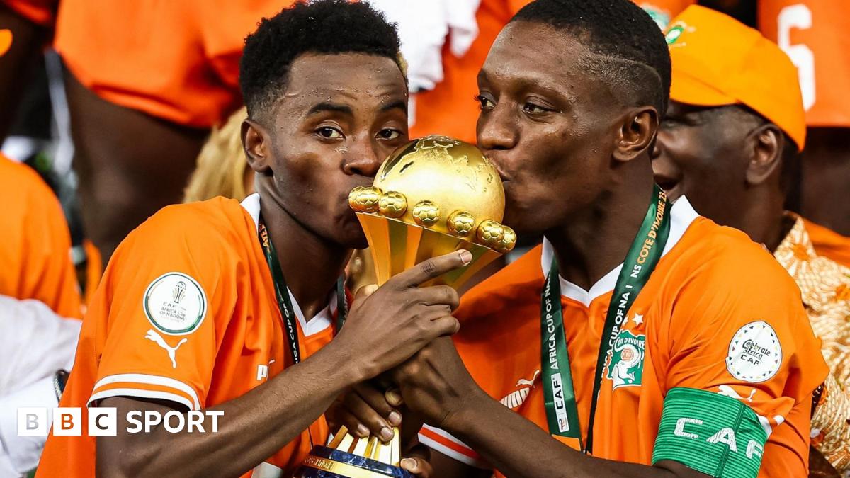 Simon Adingra and Max-Alain Gradel seen from chest up, both wearing bright orange Ivory Coast football shirts with medals around their necks, hold the golden Africa Cup of Nations trophy between them and kiss the top of it. Adingra is looking directly into the camera while Gradel casts his eyes to the left of the screen