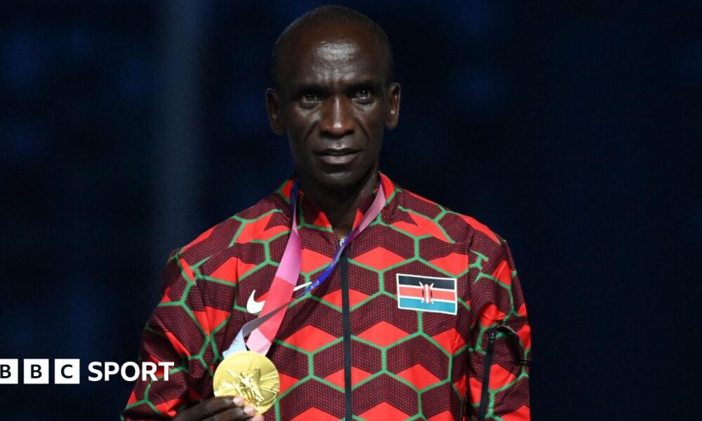 Eliud Kipchoge, visible from stomach up and wearing an official Kenya tracksuit which has the flag on its left breast and is a mainly red design with green hexagonal shapes, poses with his gold medal which he is holding in his right hand and wearing on a ribbon around his neck during the victory ceremony of the men's marathon event during the Tokyo 2020 Olympic Games