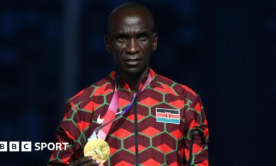 Eliud Kipchoge, visible from stomach up and wearing an official Kenya tracksuit which has the flag on its left breast and is a mainly red design with green hexagonal shapes, poses with his gold medal which he is holding in his right hand and wearing on a ribbon around his neck during the victory ceremony of the men's marathon event during the Tokyo 2020 Olympic Games