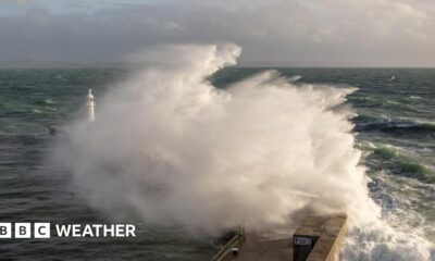 A large wave crashes against a breakwater with a lighthouse sending up a big spray of foam