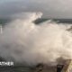 A large wave crashes against a breakwater with a lighthouse sending up a big spray of foam