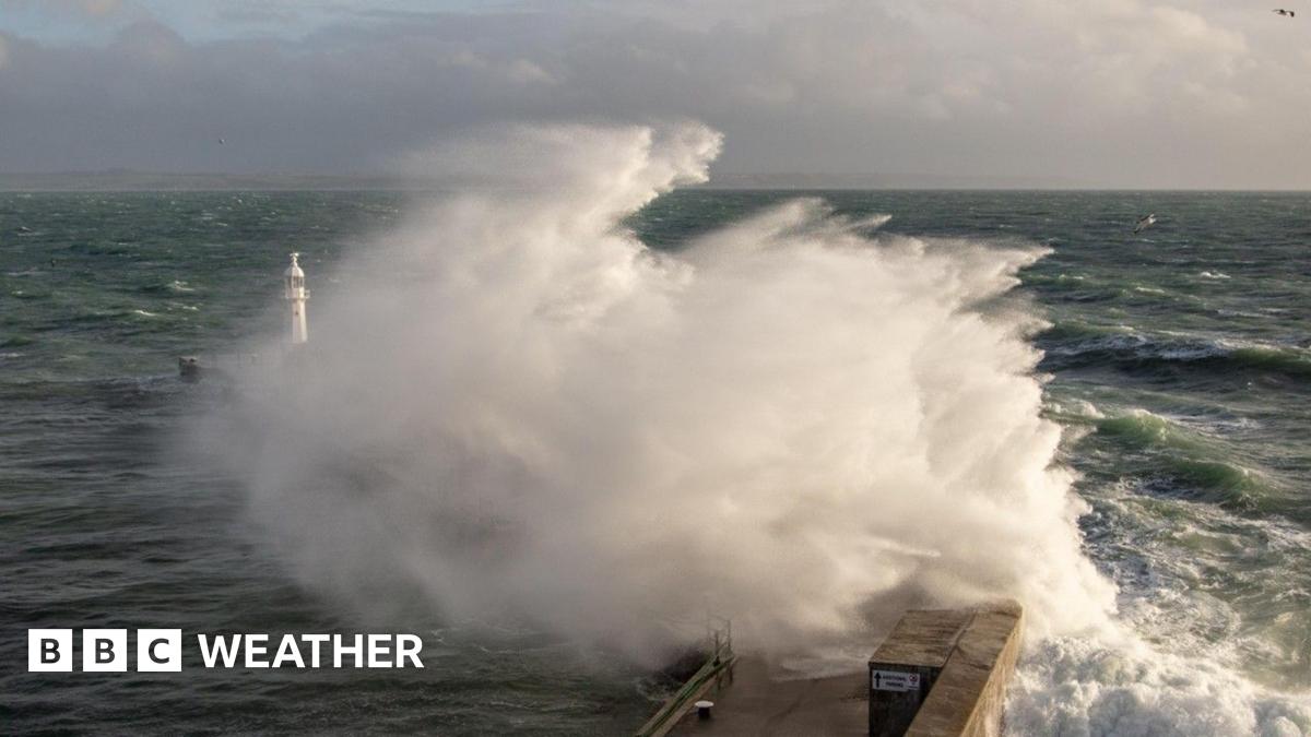 A large wave crashes against a breakwater with a lighthouse sending up a big spray of foam