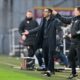 Chelsea Football Club manager Liam Rosenior standing on a sideline at a football stadium in a black outfit with his arms outstretched and other coaches either side of him in front of a green grass pitch