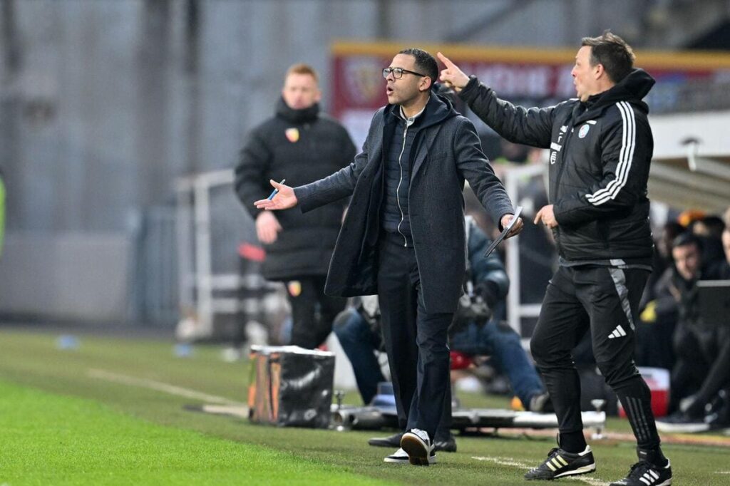 Chelsea Football Club manager Liam Rosenior standing on a sideline at a football stadium in a black outfit with his arms outstretched and other coaches either side of him in front of a green grass pitch