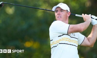 Rory McIlroy watches his ball after hitting a shot with a wood. He is wearing a European Ryder Cup outfit - a baseball cap and a predominantly white polo shirt with black and yellow narrow hoops and trim.