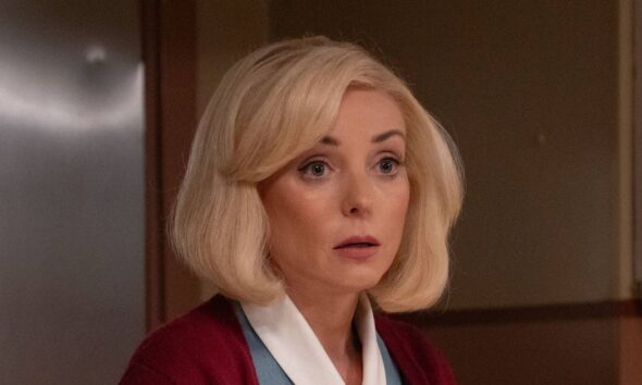 An individual in formal attire, accompanied by another person dressed in a blue gown, stands on a balcony, their gaze directed towards a body of water with boats and distant structures visible in the background.