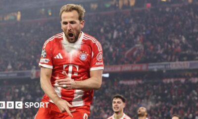 Harry Kane celebrates a goal for Bayern Munich, with his mouth wide open. He is wearing Bayern's largely red home kit, with white stripes on the sleeves, and a white collar and cuffs, with large crowd visible in the background