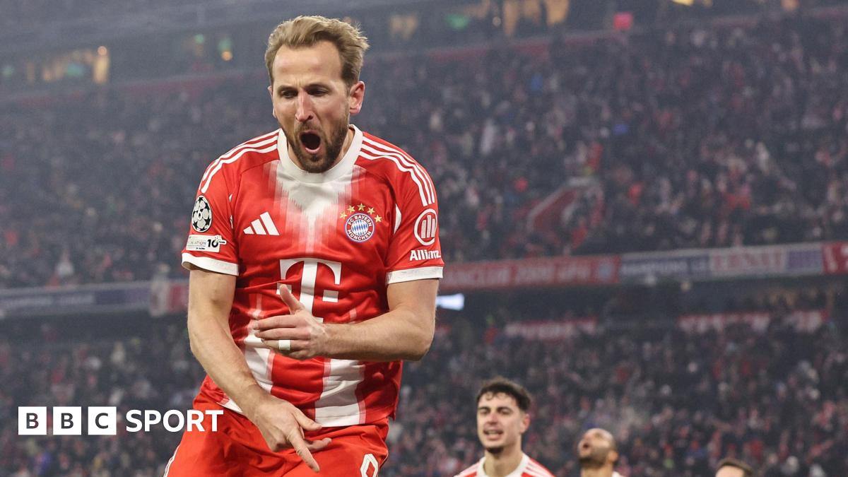 Harry Kane celebrates a goal for Bayern Munich, with his mouth wide open. He is wearing Bayern's largely red home kit, with white stripes on the sleeves, and a white collar and cuffs, with large crowd visible in the background