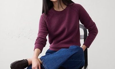 A woman with a purple sweater, blue jeans, and brown loafers. She is sitting on a black chair with her legs crossed.