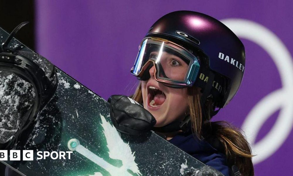 Mia Brookes shows an expression of delight, with her mouth wide open as she wears a protective dark-coloured helmet and ski goggles while carrying a snowboard