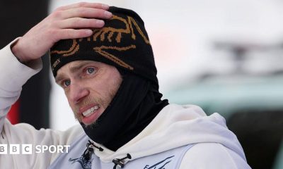 Gus Kenworthy is wearing a black beanie and snood and has his hand on his head