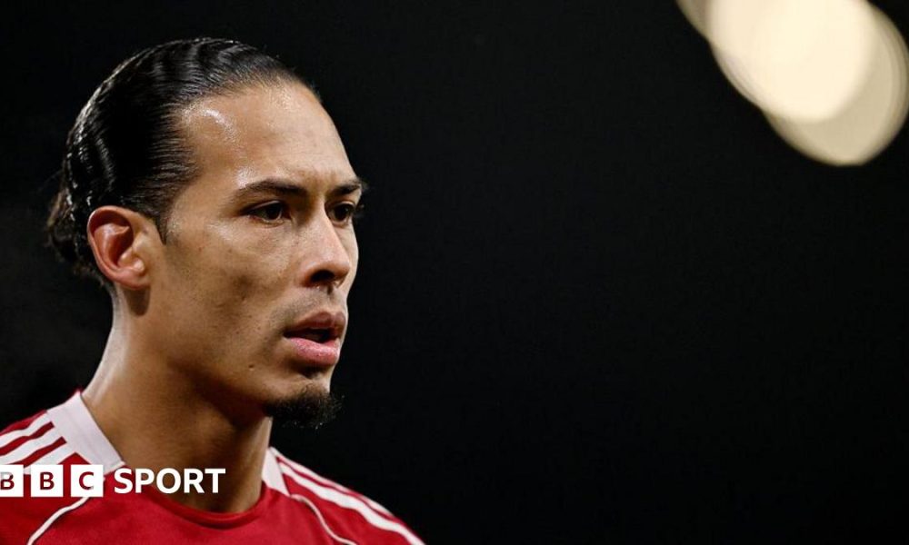 Close-up image of Virgil van Dijk in the left of the photograph. He has black hair, tied back, and is wearing Liverpool's red home shirt. The background of the image is black with a spotlight in the top right corner.