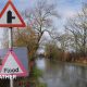 A flooded road flanked by trees with a blue and grey sky overhead and a sign in the foreground that reads "Flood".