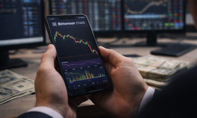 An investor holds a smartphone displaying a cryptocurrency candlestick chart while monitoring markets at a trading desk.