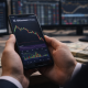 An investor holds a smartphone displaying a cryptocurrency candlestick chart while monitoring markets at a trading desk.