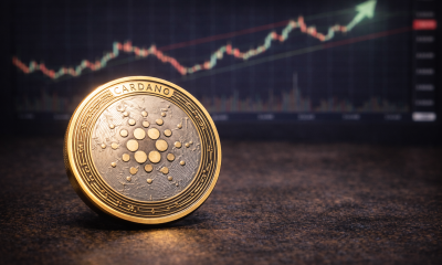 A Cardano (ADA) cryptocurrency token placed on a table with a blurred upward-trending market chart in the background.