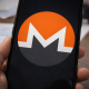 A smartphone held in hand displays the Monero cryptocurrency logo on its screen, with financial documents blurred in the background.