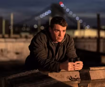 Gabriel Basso as Peter Sutherland leaning on a wall outside in Season 2 of The Night Agent