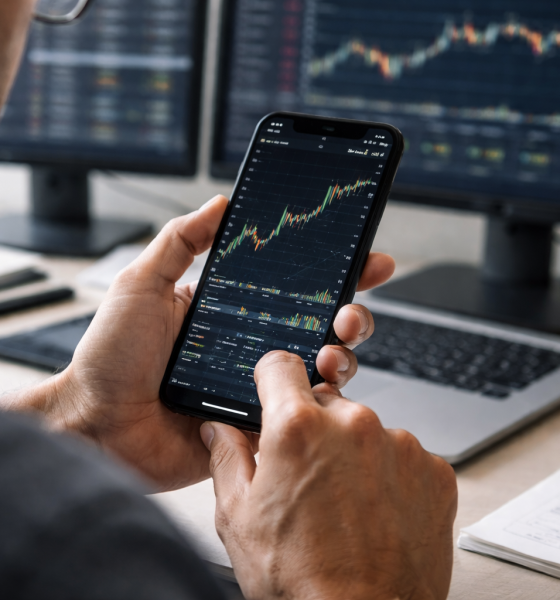 A trader analyzes a financial price chart on a smartphone while multiple market charts display on monitors in the background.