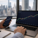 An investor checks rising cryptocurrency charts on a laptop and smartphone with a city skyline visible through the office window.