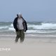 Man and his dog walking along a beach where sand is visibily lifted up with large waves in the background