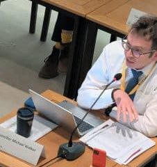 New Reform UK councillor, Michael Walker sat alone at a table. He has a laptop and a phone propped upright and some papers in front of him. He is gesturing with and open downward palm and he has his mouth open as he speaks.